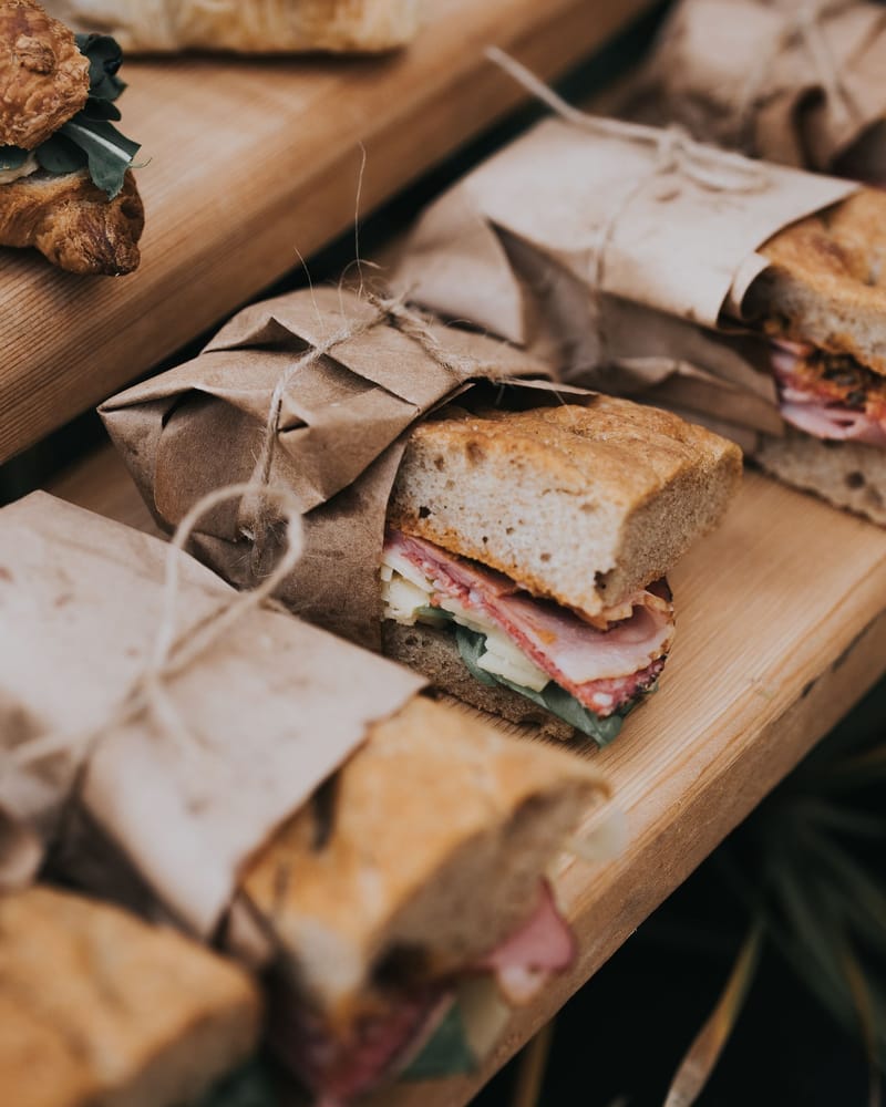 Italian sandwiches on focaccia, each half-wrapped in kraft paper and tied with twine, resting on pale wooden boards.